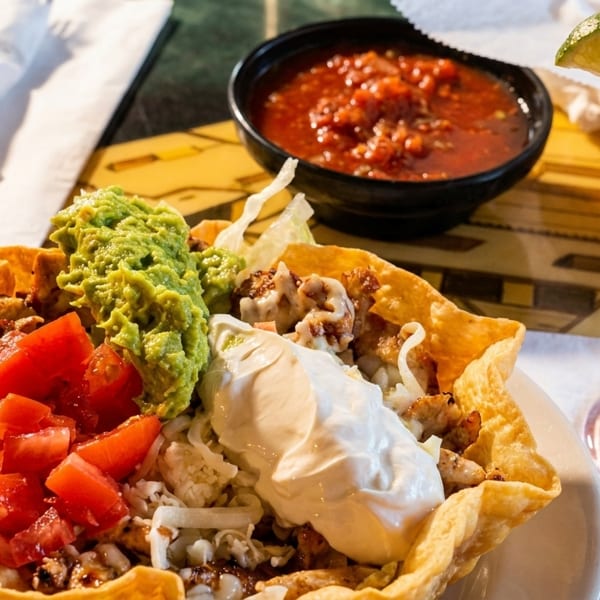 A taco salad in a crispy shell with guacamole, tomatoes, and sour cream, next to salsa and a red margarita.