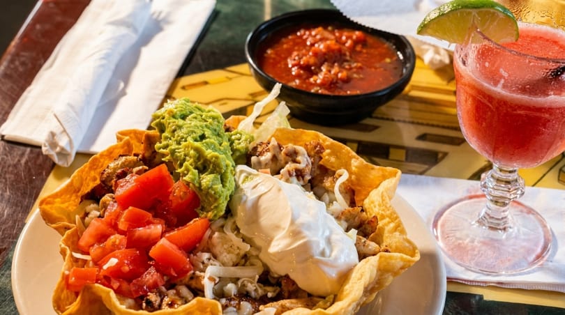 A taco salad in a crispy shell with guacamole, tomatoes, and sour cream, next to salsa and a red margarita.