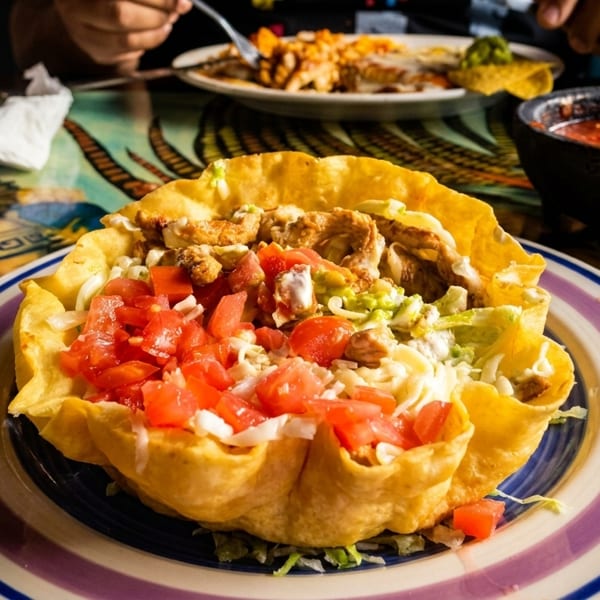 A chicken taco salad in a crispy tortilla bowl with diced tomatoes on a colorful table.