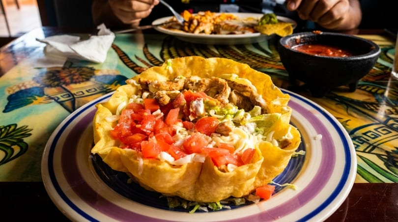 A chicken taco salad in a crispy tortilla bowl with diced tomatoes on a colorful table.