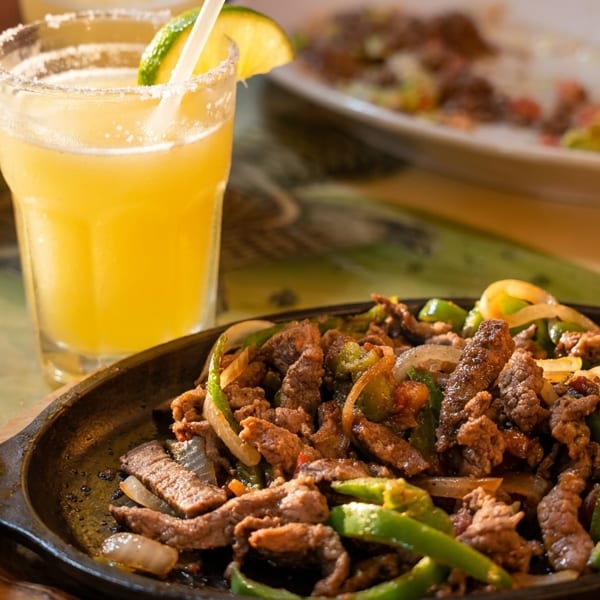 Sizzling beef fajitas with green peppers and onions, next to a margarita with a lime.