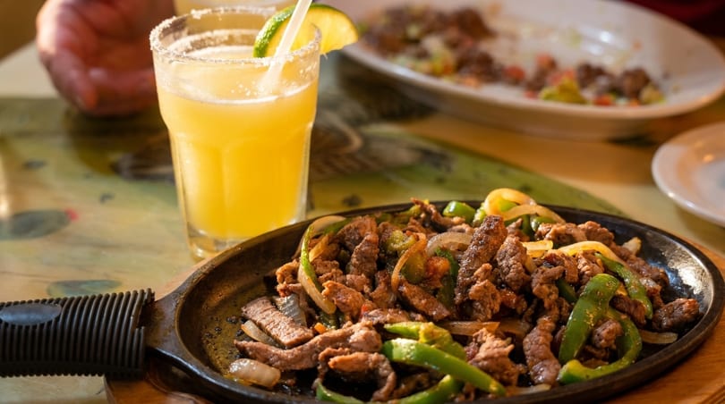 Sizzling beef fajitas with green peppers and onions, next to a margarita with a lime.