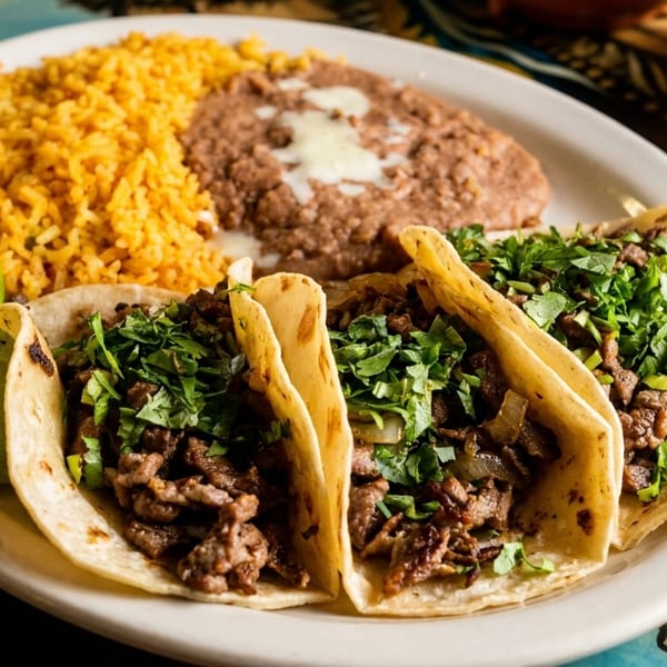 Three carne asada tacos with cilantro, rice, refried beans, and lime wedges on a white plate.