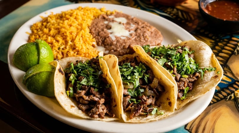 Three carne asada tacos with cilantro, rice, refried beans, and lime wedges on a white plate.