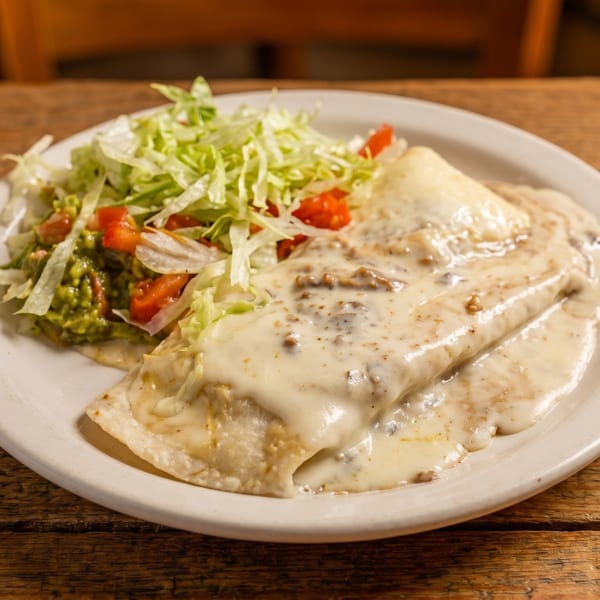 Burrito covered in white sauce, with a side of guacamole, lettuce, and diced tomatoes on a wooden table.
