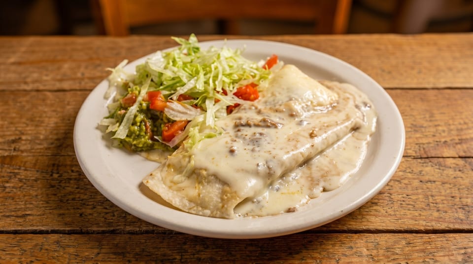 Burrito covered in white sauce, with a side of guacamole, lettuce, and diced tomatoes on a wooden table.